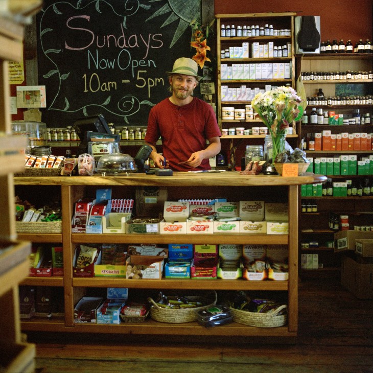 Man in red shirt and green hat stands behind counter at organic food shop with shelves of products and chalkboard sign