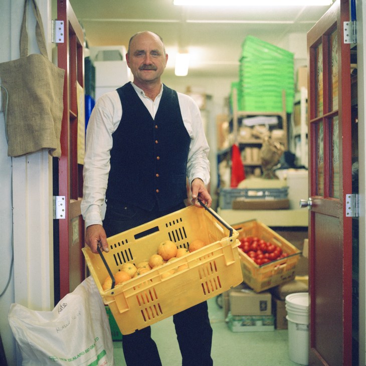 Shop manager in dark vest and white shirt holding a yellow crate of fresh produce in a storage corridor.
