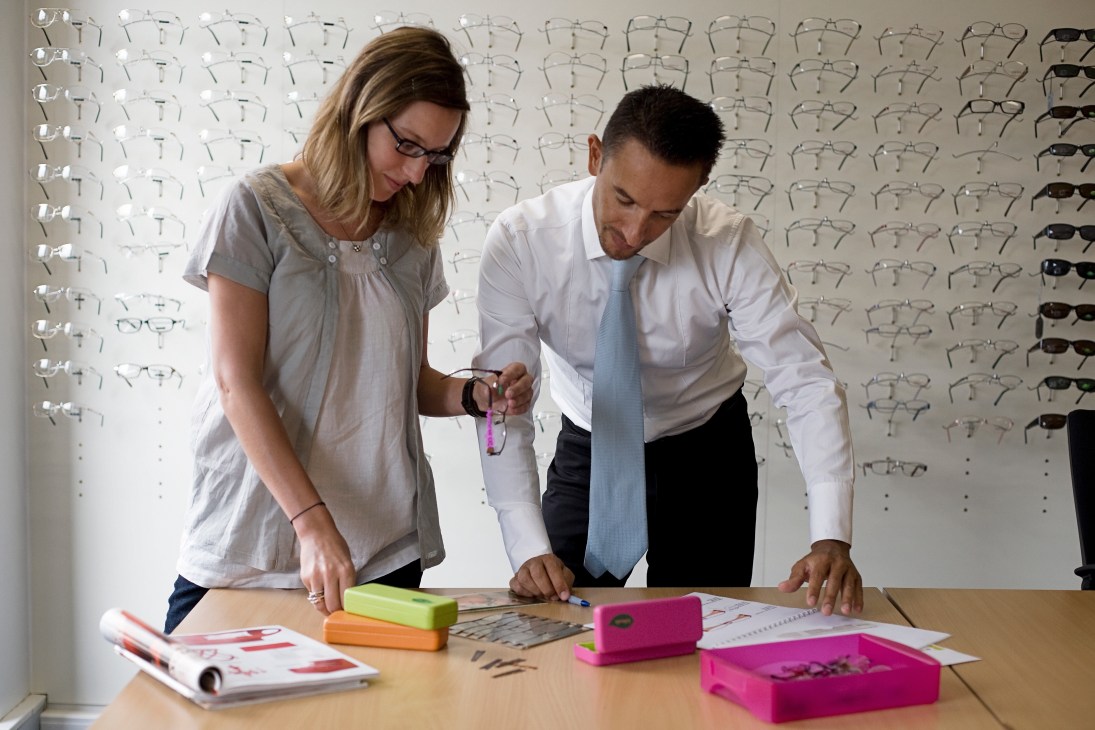 Two Atol employees review spectacle frame designs at a table with samples and sketches, with eyeglass frames displayed on the wall behind them.