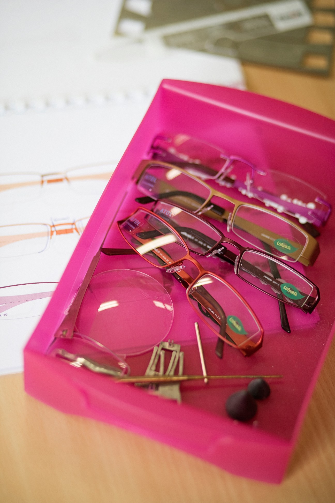 Multiple eyeglass frames in various styles displayed in a bright pink container on a wooden surface.