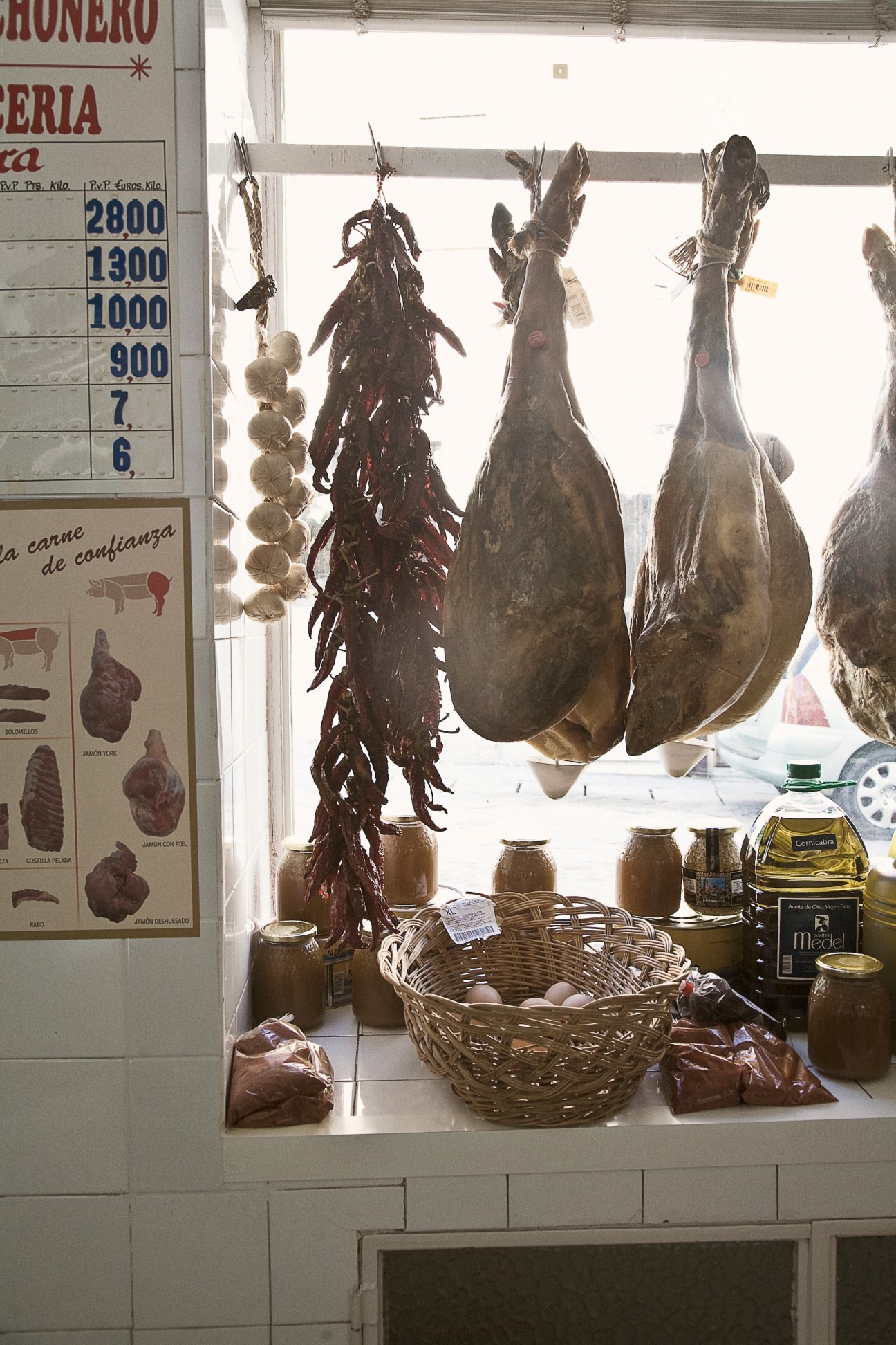 Cured hams and chorizo hanging in a butcher shop window with price list and olive oil bottles.