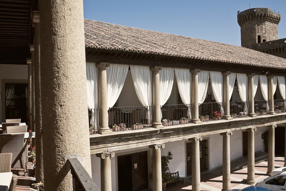 Historic stone building with columned arcade and white curtains overlooking a courtyard with a tower in the background.