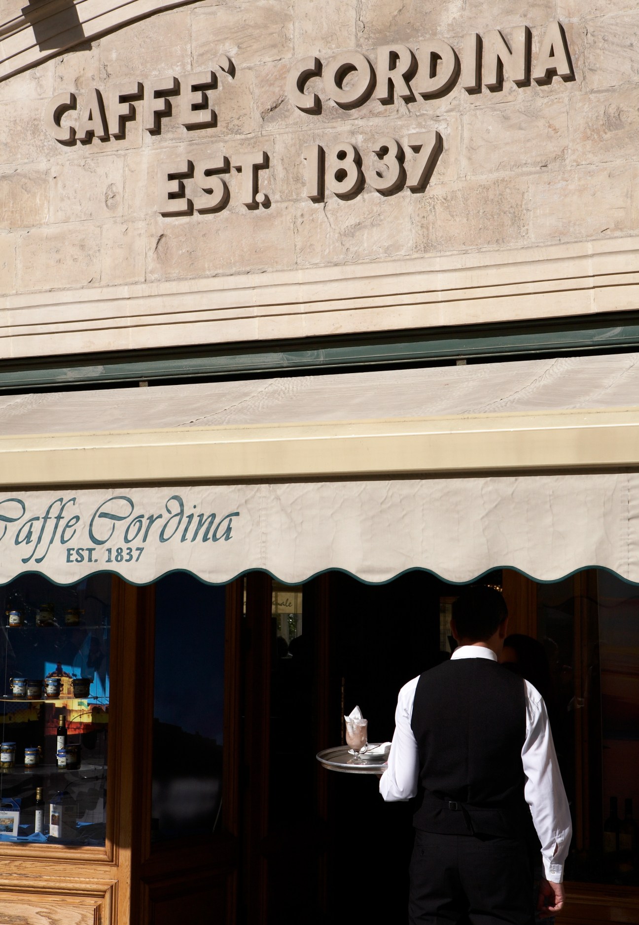 Caffe Cordina storefront with 1837 establishment date and waiter carrying tray outside.