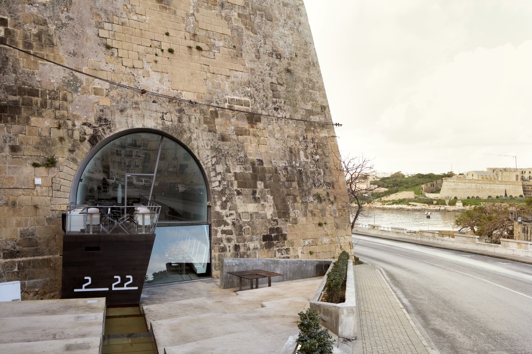 Modern bar entrance with circular glass window built into historic fortress stone wall overlooking Grand Harbour.