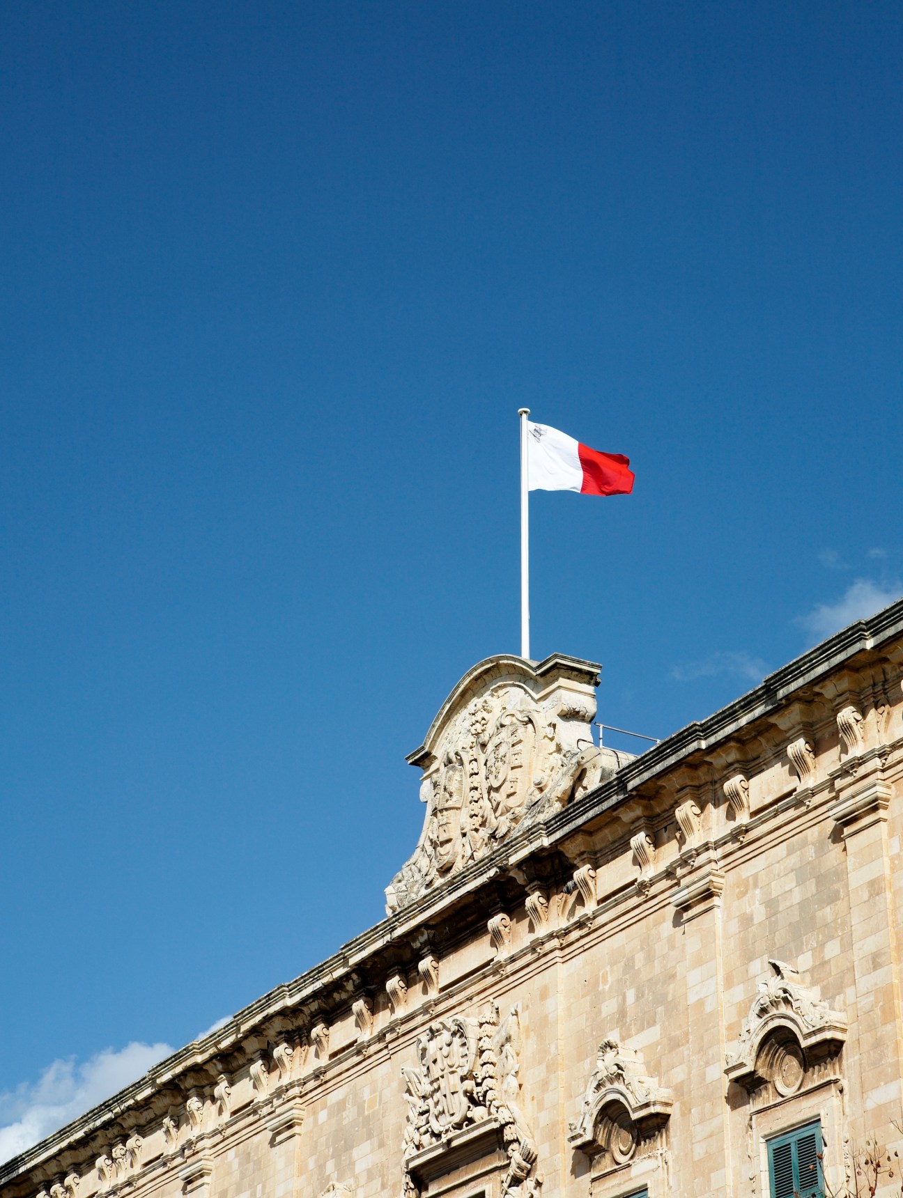 Maltese flag flying above ornate stone building with carved architectural details.