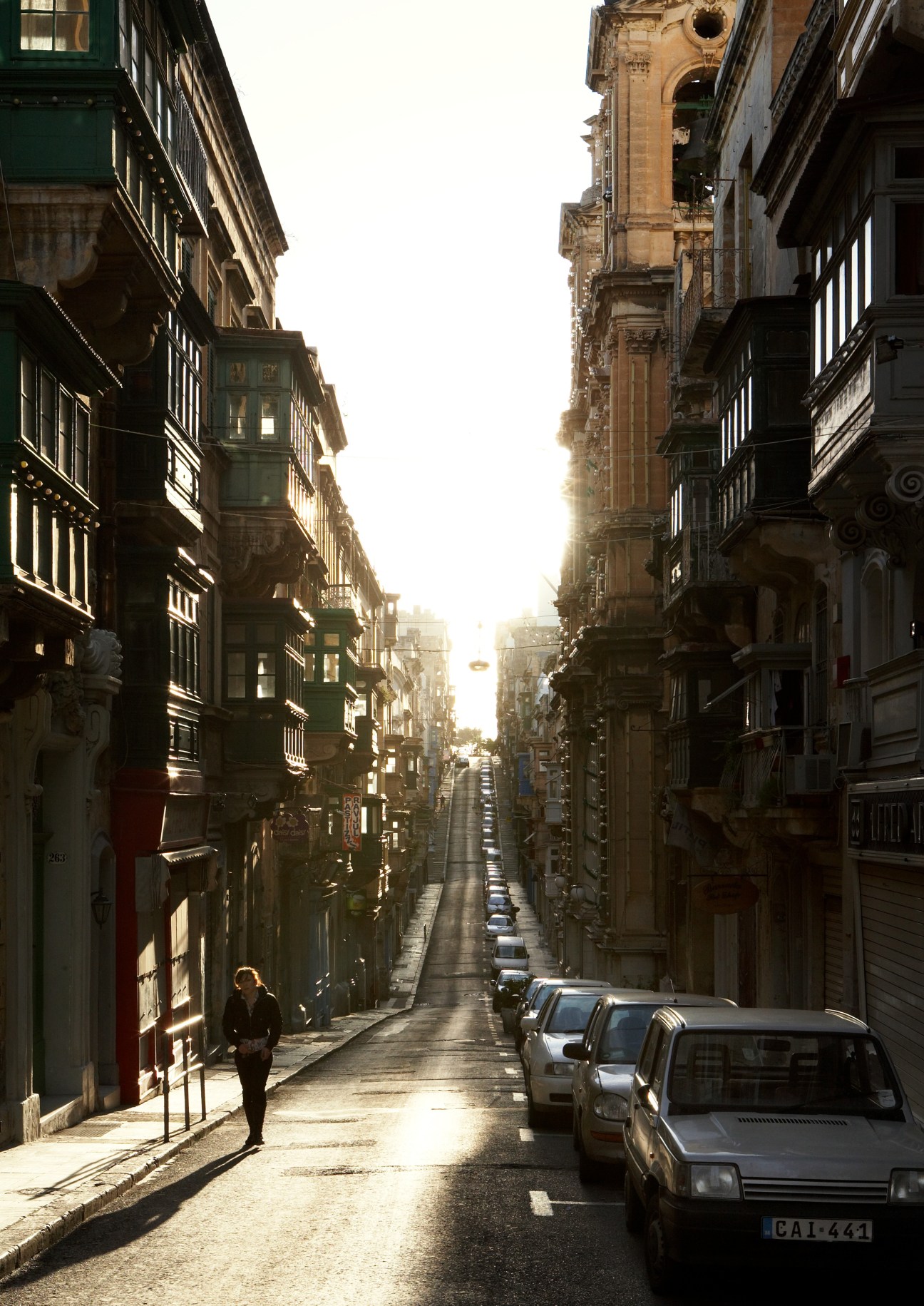 Narrow sunlit street in Valletta's old town with historic stone buildings and parked cars