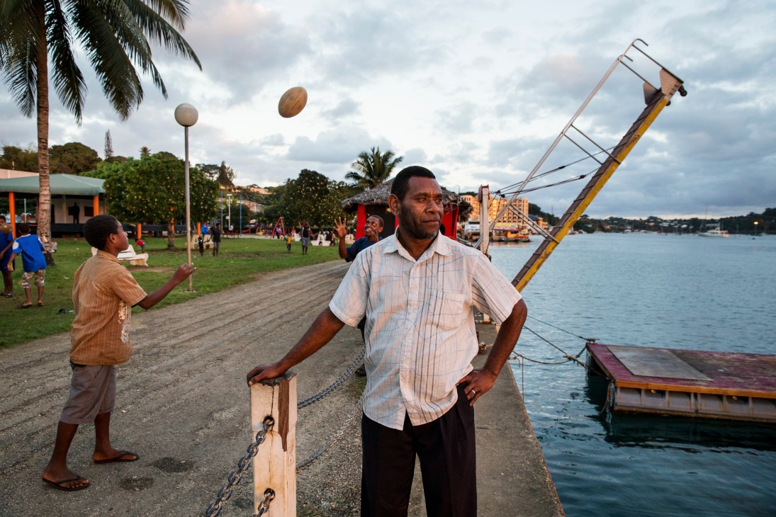 A man in a light pink shirt stands on Port Vila's waterfront with boats and palm trees visible behind him.