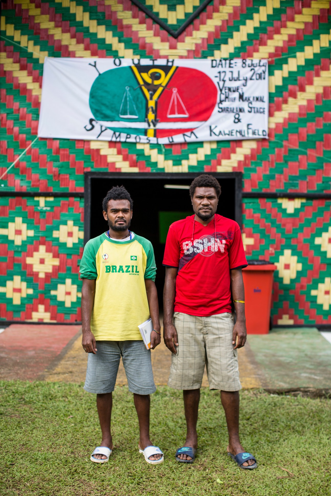 Two young men stand in front of a colorful woven building backdrop with a youth justice conference banner.