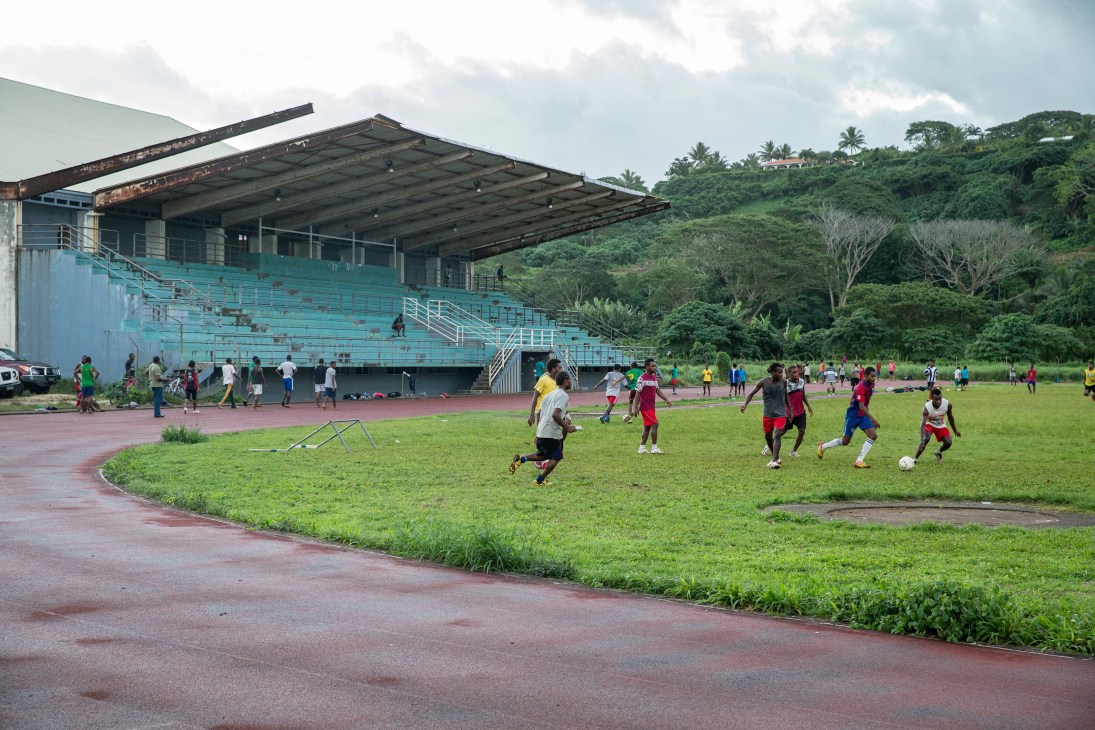 Children playing soccer on a grass field in front of a deteriorating turquoise stadium with rusted metal roof in Port Vila, Vanuatu.
