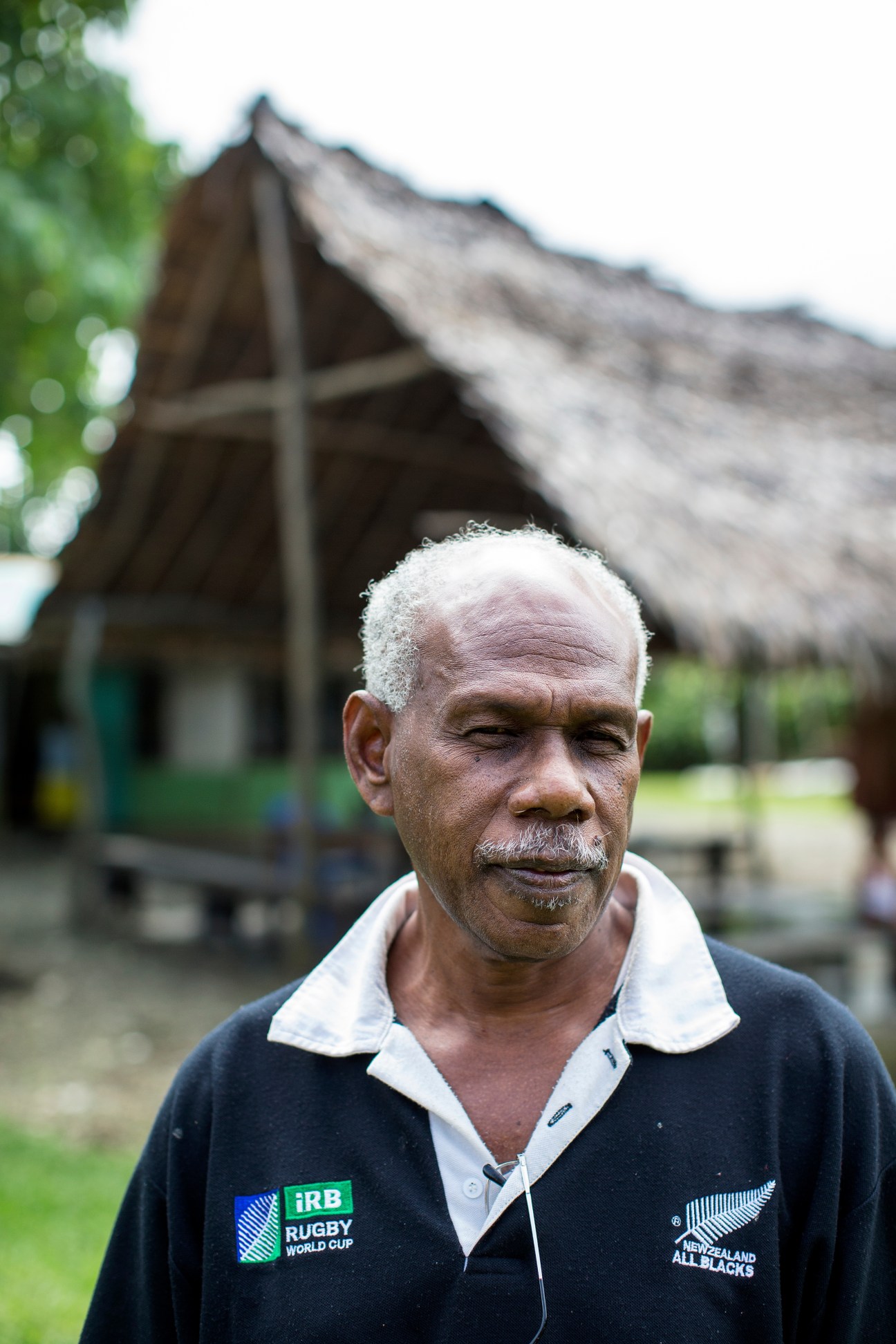Peter Kalmos Kalontan, chairman of the Council of Chiefs in Pango, standing in front of a traditional thatched-roof building.
