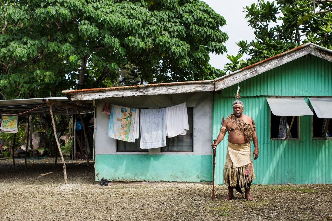 Chief Roland Kaluatman Maseiman stands outside a green corrugated metal house in traditional dress.