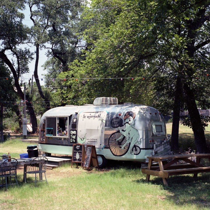 Vegan Yacht food trailer with illustrated cyclist artwork parked under trees with picnic seating.