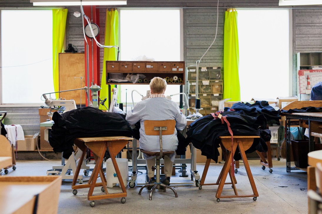 Seamstress working on black garments at wooden tailoring tables in a bright factory workshop with lime-green curtains.