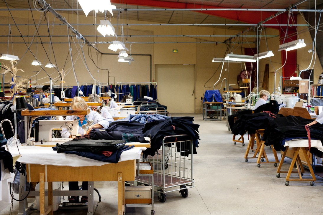 Workers sewing dark navy and black clothing at wooden tables in a bright factory floor with hanging lights and fabric racks.