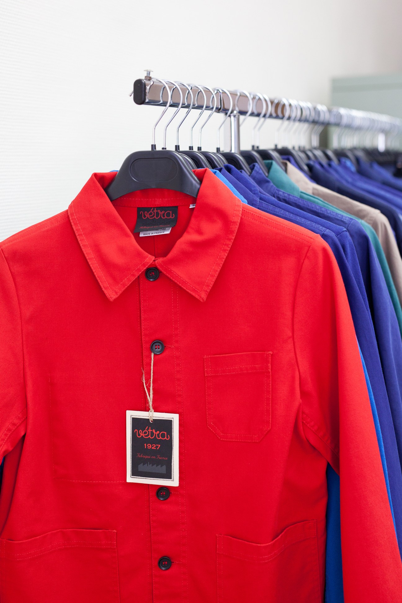 Vétra workwear jackets in bright red, blue, and other colors hanging on a rail, with a label reading 'vétra 1927'.