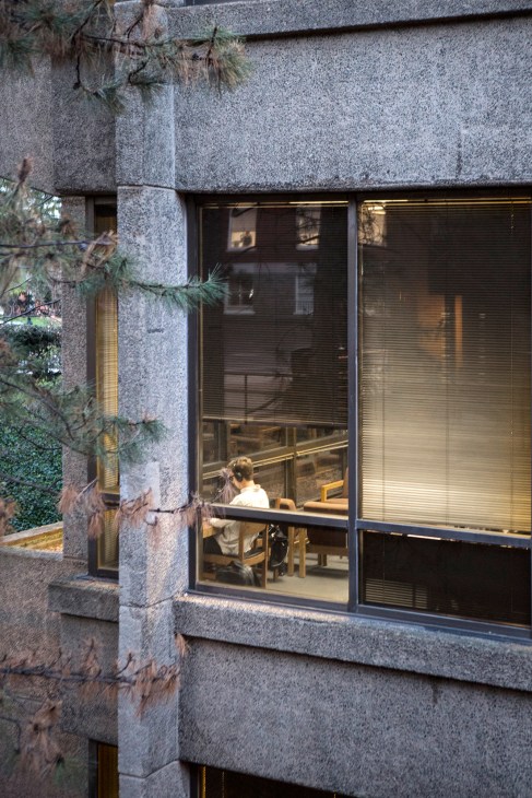 A student studies at a desk visible through large windows of a brutalist concrete building.