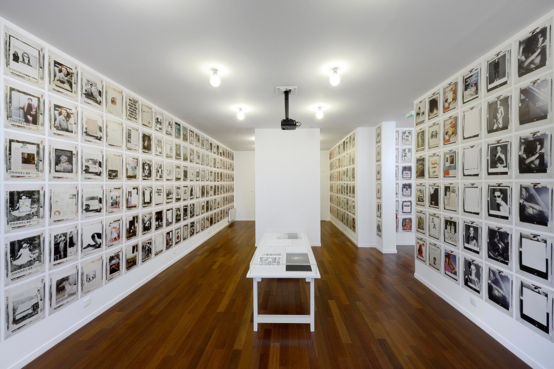 Gallery exhibition space with framed artworks covering two walls and a central display table.