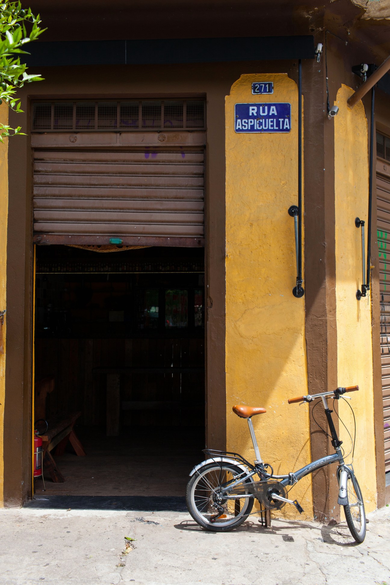 Folding bicycle leaning against a bright yellow building with a blue street sign reading Rua Aspicuelta on a residential street.