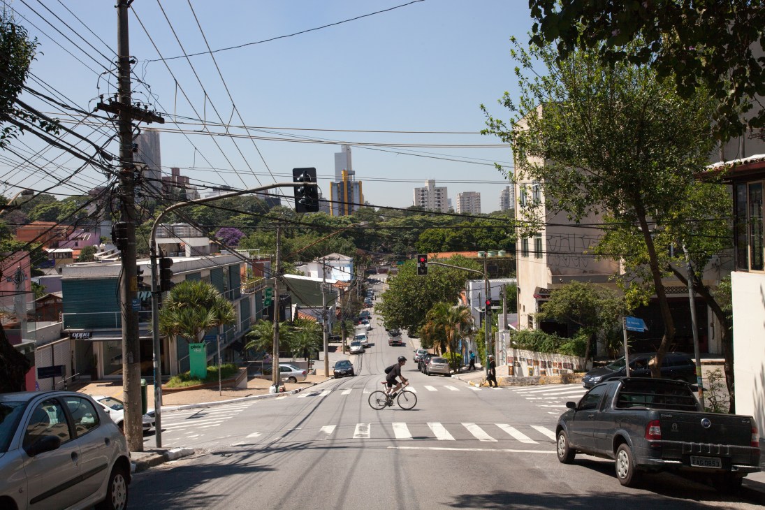 A tree-lined street in Vila Madalena with low-rise buildings, a cyclist crossing at a traffic light, and São Paulo's skyline visible in the distance.