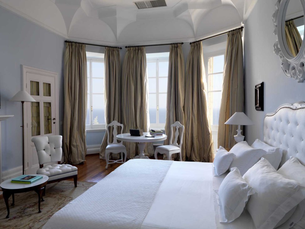 Elegant bedroom with white upholstered bed, curved windows with gold curtains, and sea view at Villa della Pergola.