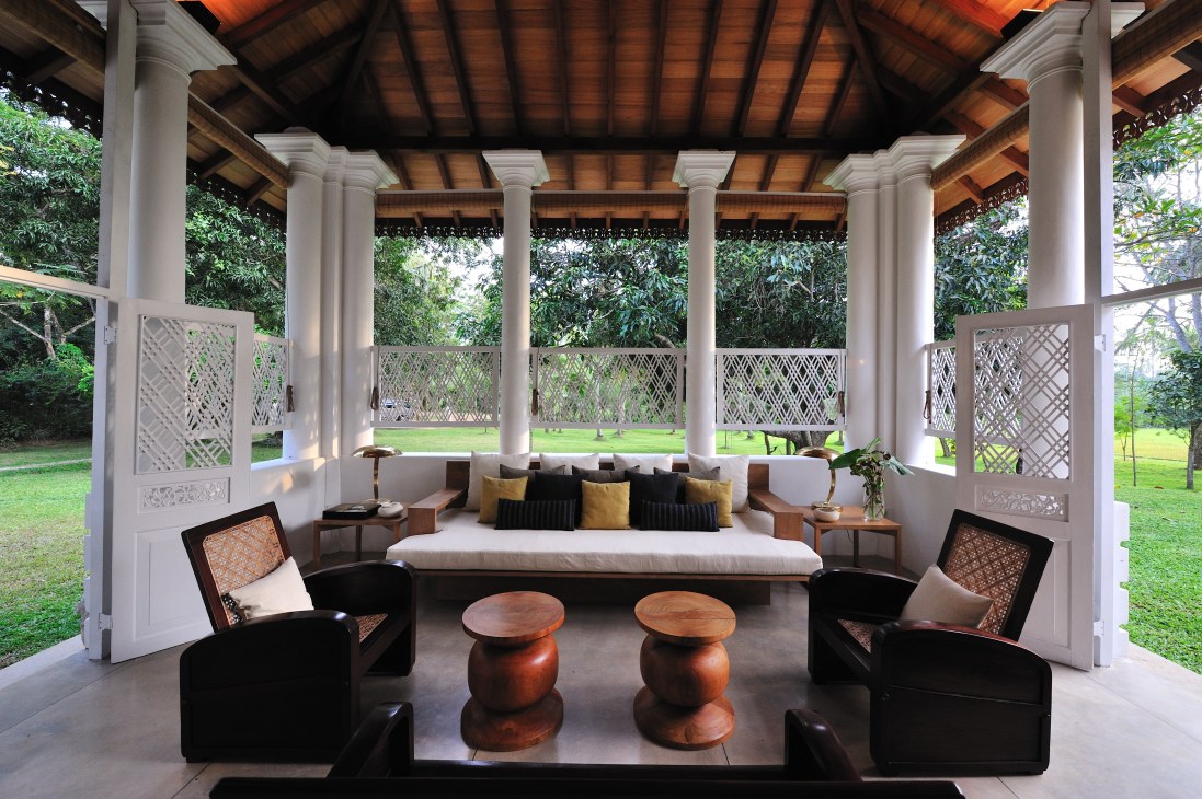 Open-air pavilion with white columns, wooden ceiling, black armchairs and wooden side tables overlooking manicured lawn.