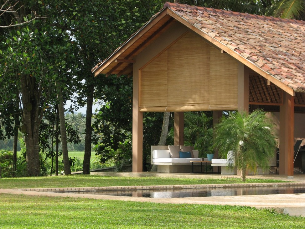 A modern pavilion with terra cotta roof and wooden shutters overlooks a pond in lush tropical gardens surrounded by tall trees.
