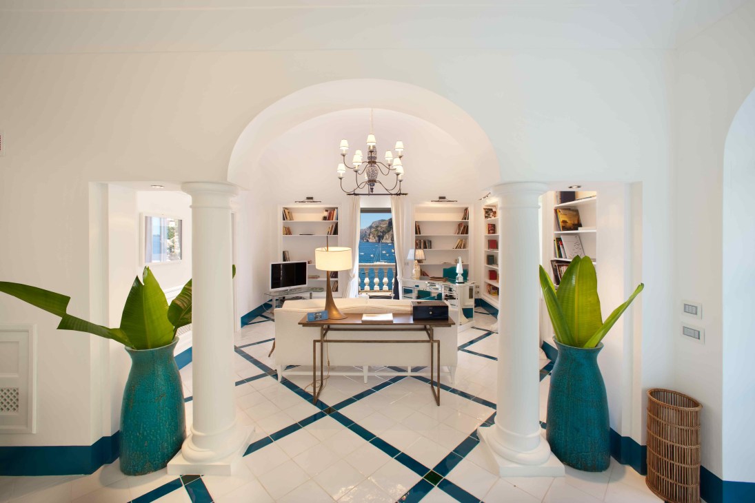 Bright white interior room with arched ceiling, blue geometric floor pattern, teal planters with green plants, white bookshelves, and sea view through central doorway.