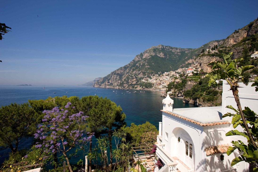 White villa with terracotta roof overlooking the Amalfi Coast with purple flowering plants and boats in the blue sea.