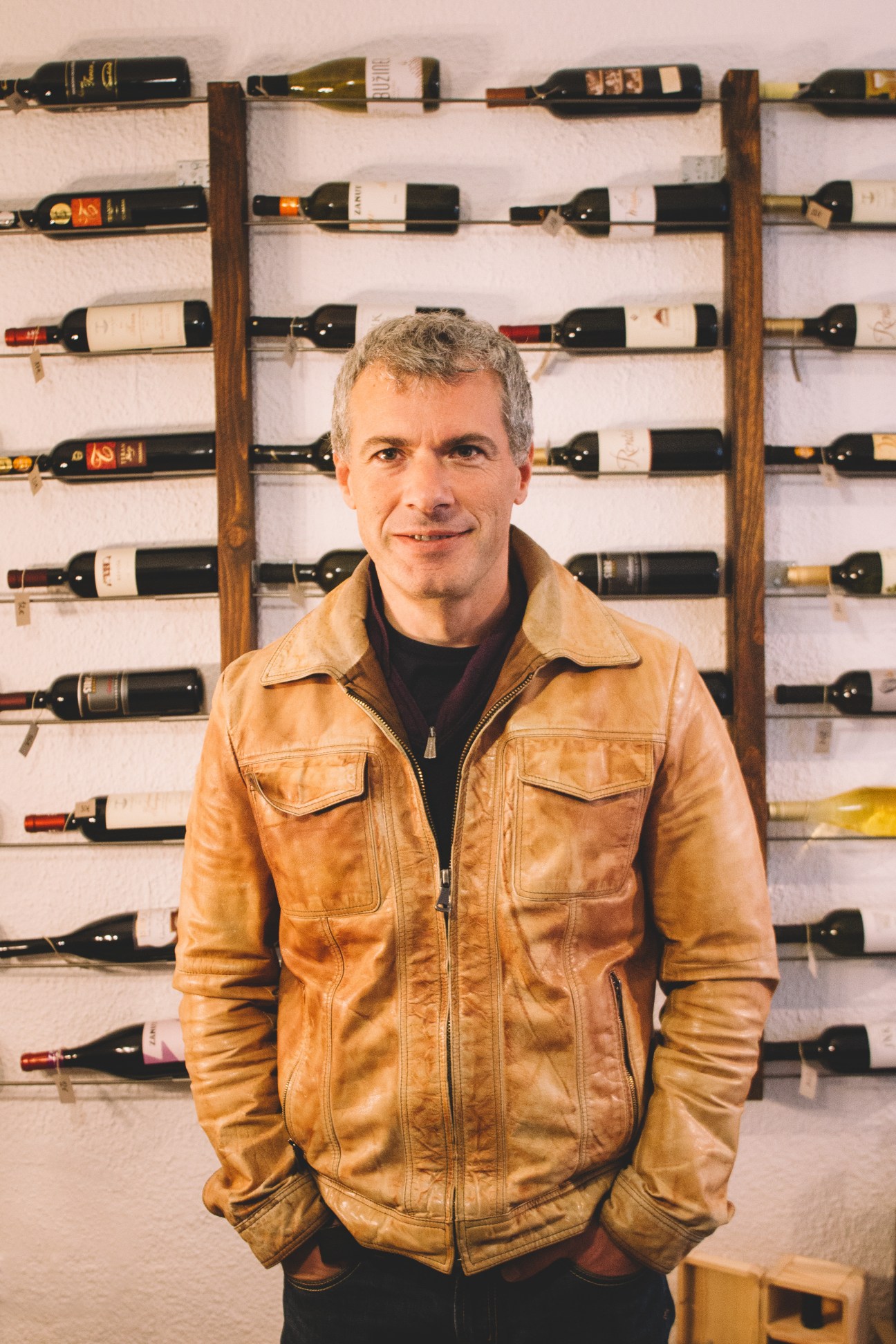 Man in tan leather jacket standing in front of wine bottles displayed on wooden wall racks