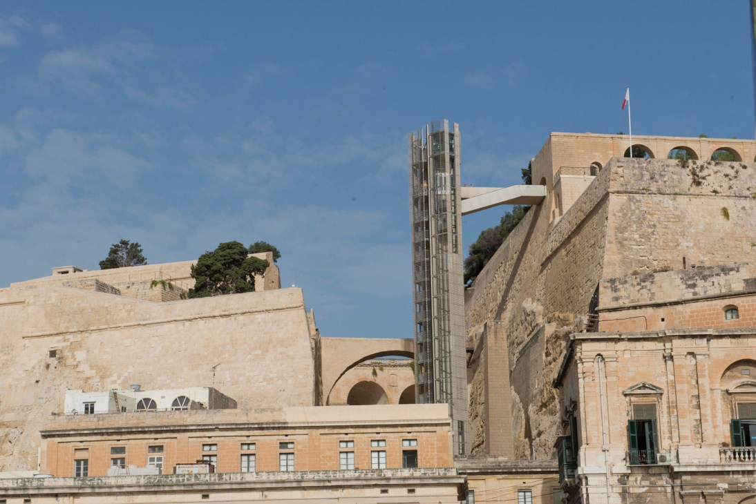The Barrakka lift, a modern glass elevator structure connecting Valletta's lower and upper levels, surrounded by historic stone fortifications and buildings.