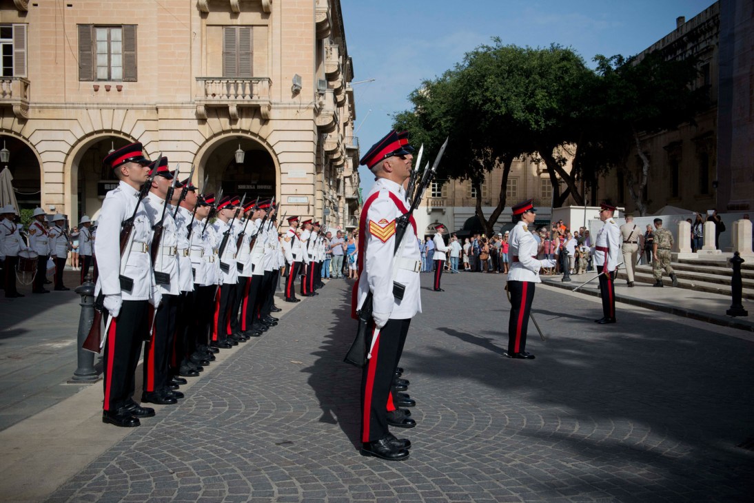 Maltese soldiers in white uniforms with red and black stripes standing in formation in a historic square.