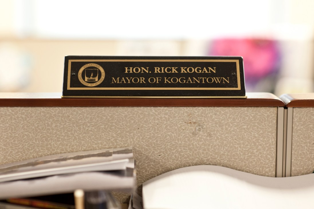 A nameplate reading 'Hon. Rick Kogan Mayor of Kogantown' sits on a desk with papers nearby.