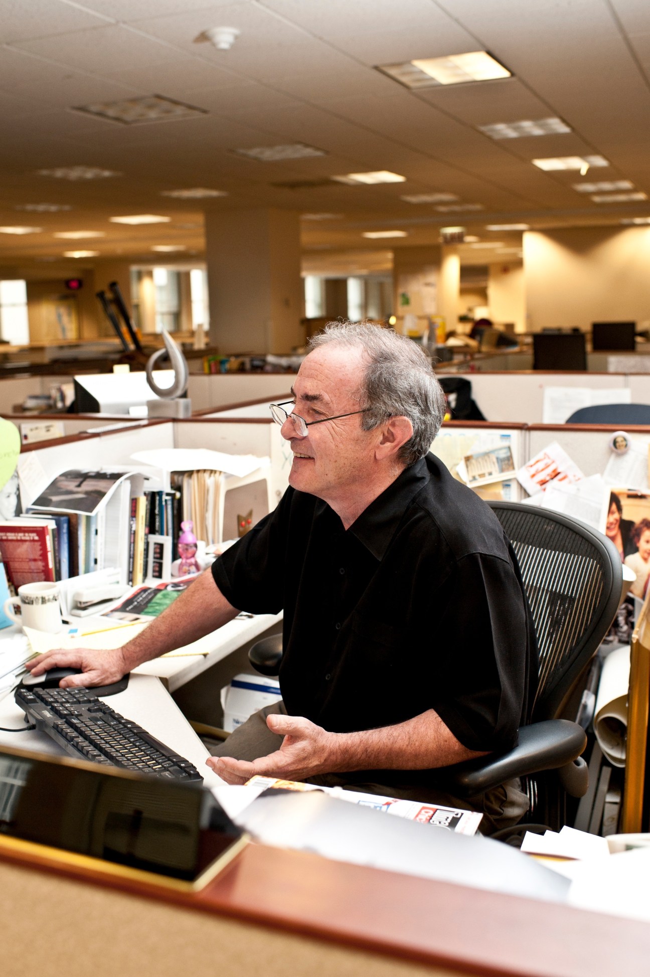 Rick Kogan sits at his desk in the Chicago Tribune newsroom, wearing glasses and a black shirt.