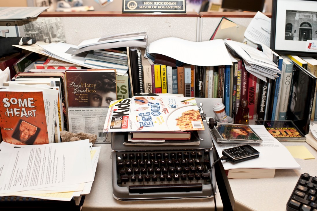 Rick Kogan's cluttered desk at the Chicago Tribune featuring a typewriter surrounded by books, magazines, and papers with a nameplate reading "Hon. Rick Kogan Mayor of Kogantown" visible in the background.