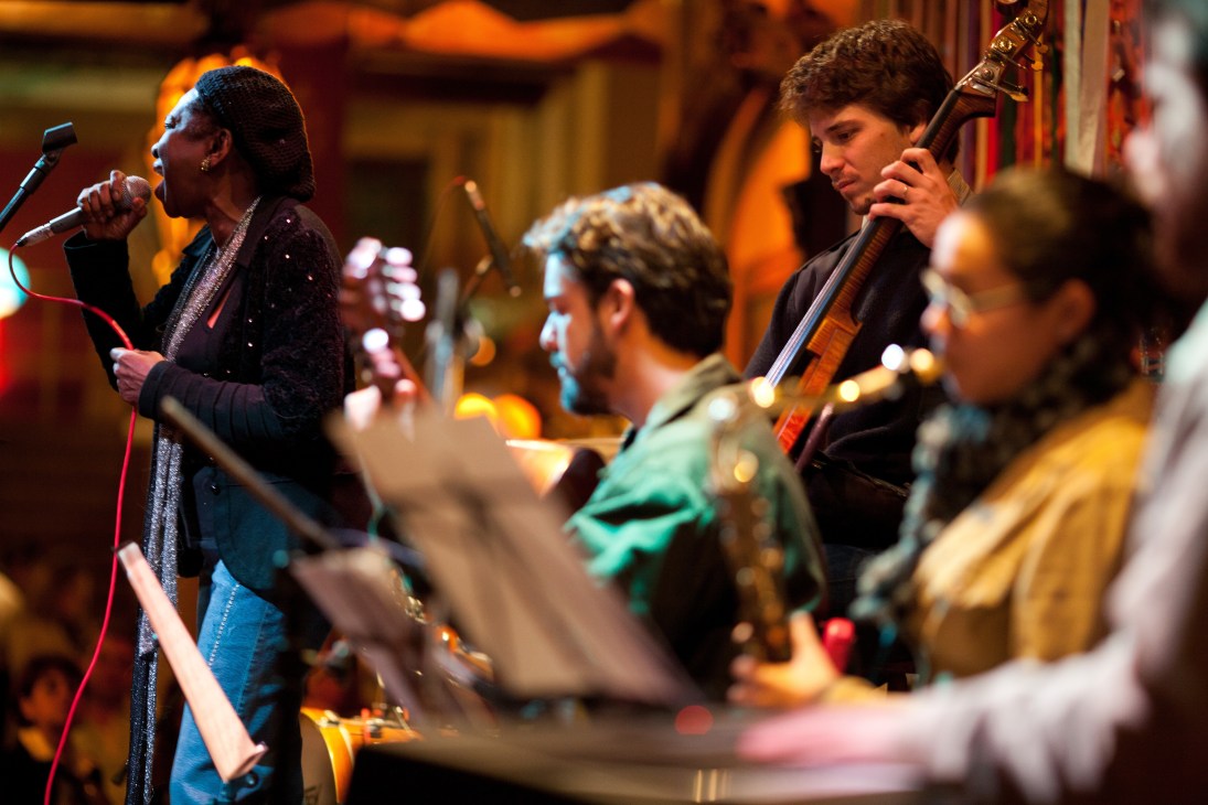 Áurea Martins performs samba at Rio Scenarium, singing into a microphone with musicians playing instruments behind her.