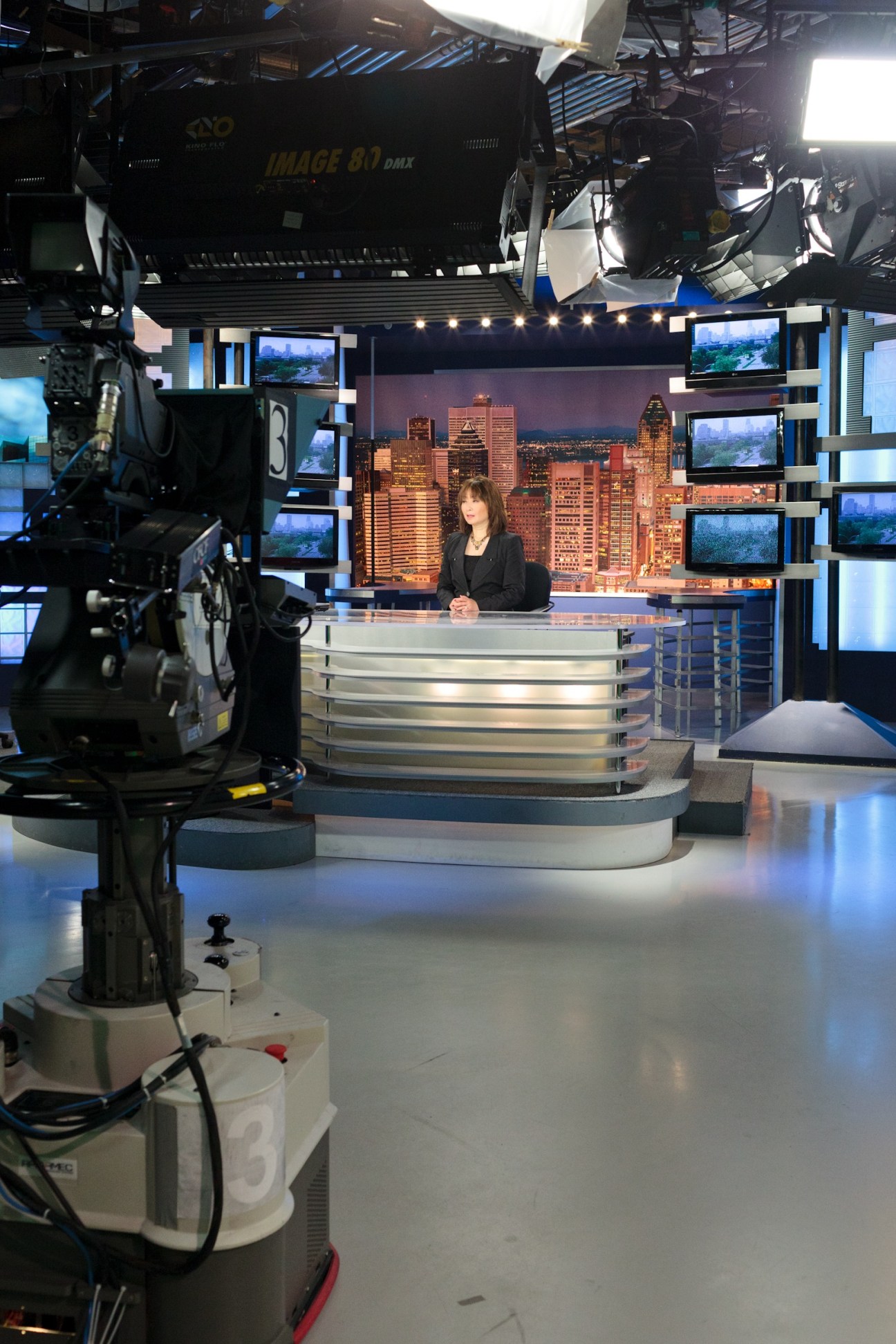 Woman in black blazer sits at news desk in TV studio with Chicago skyline backdrop and cameras overhead.