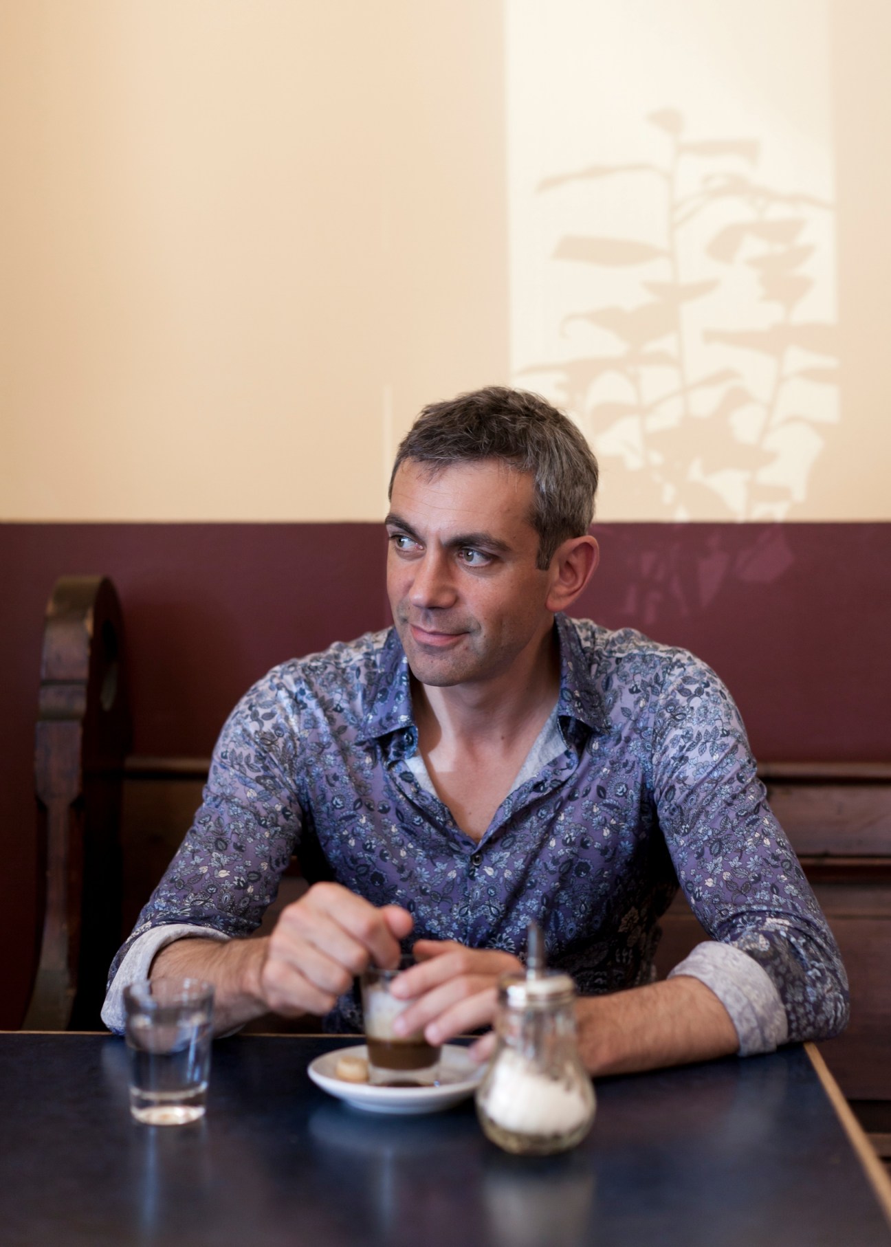 Wladimir Kaminer sits at a table in Café Meta, gesturing while speaking, with a glass of water and plate in front of him.