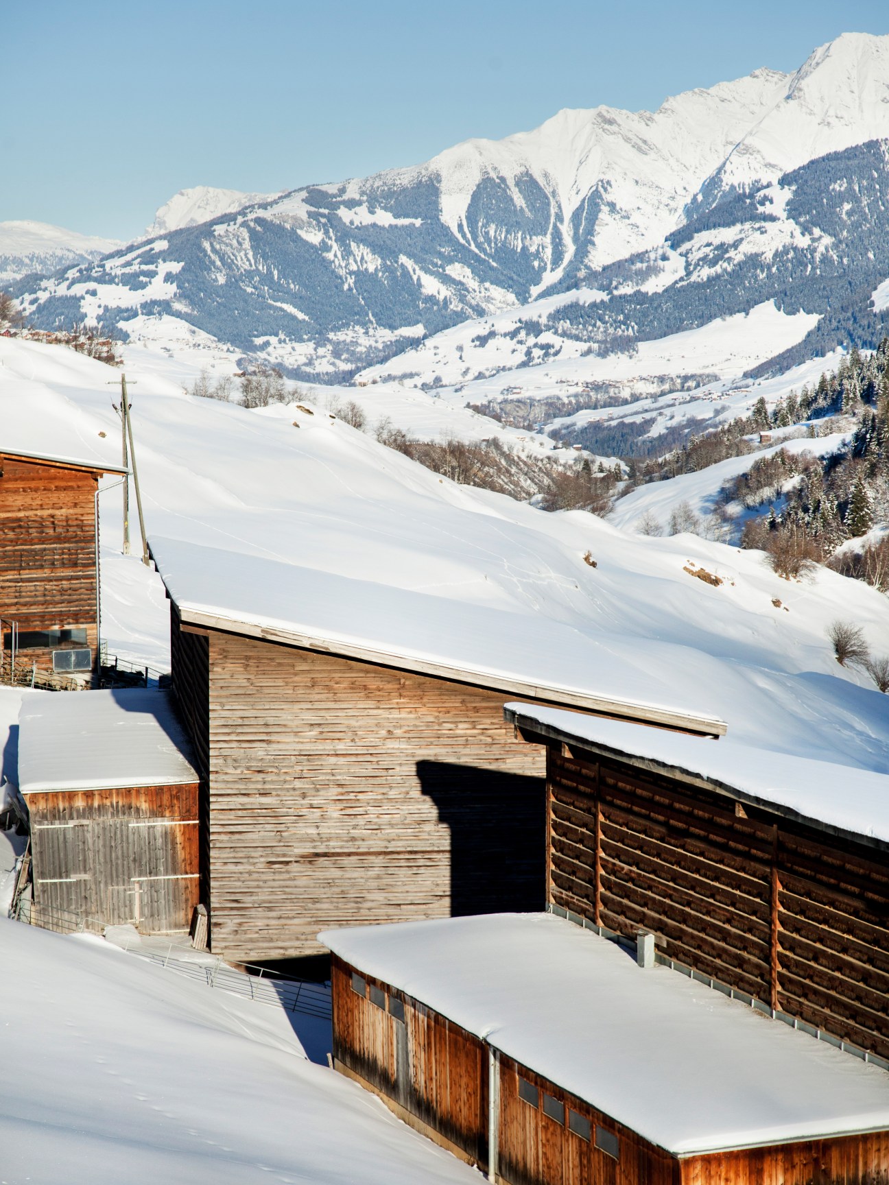 Wooden barns with snow-covered roofs nestled in a snowy Alpine valley with mountains in the background.