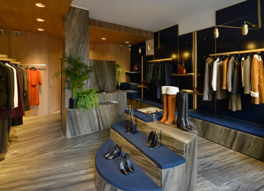 Vanessa Seward boutique interior featuring elm wood panels, clothing displays on brass rods, boots on blue pedestals, and brass pendant lights.
