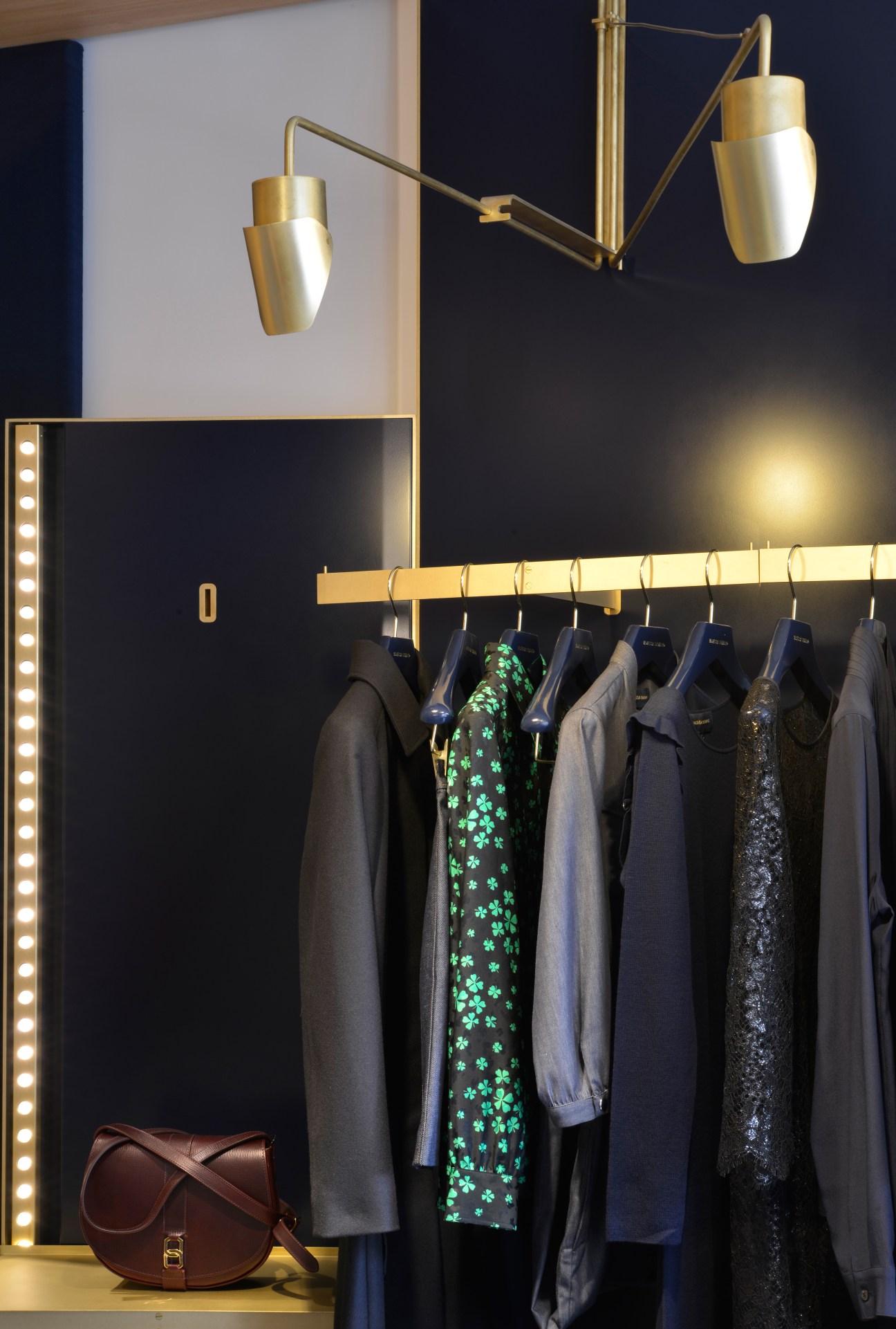Vanessa Seward boutique window displaying clothing on a brass rod with brass pendant lights and a brown leather bag.
