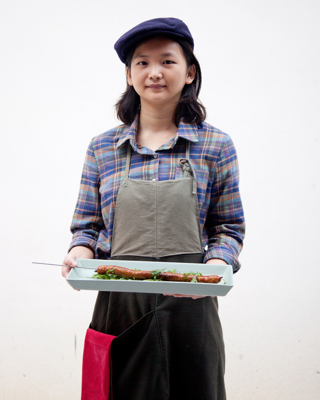 Young woman wearing a navy beret and plaid shirt holds a white plate with grilled sausages and greens.