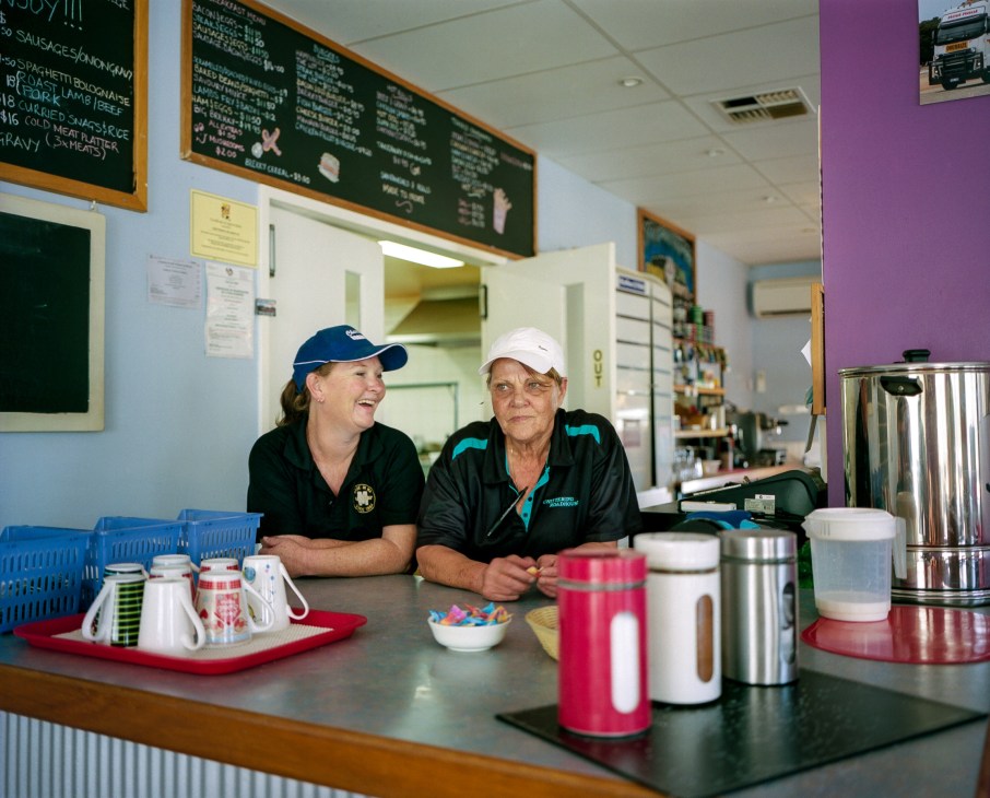 Two women smiling behind the counter at Chittering roadhouse, with menu boards and kitchen equipment visible behind them.