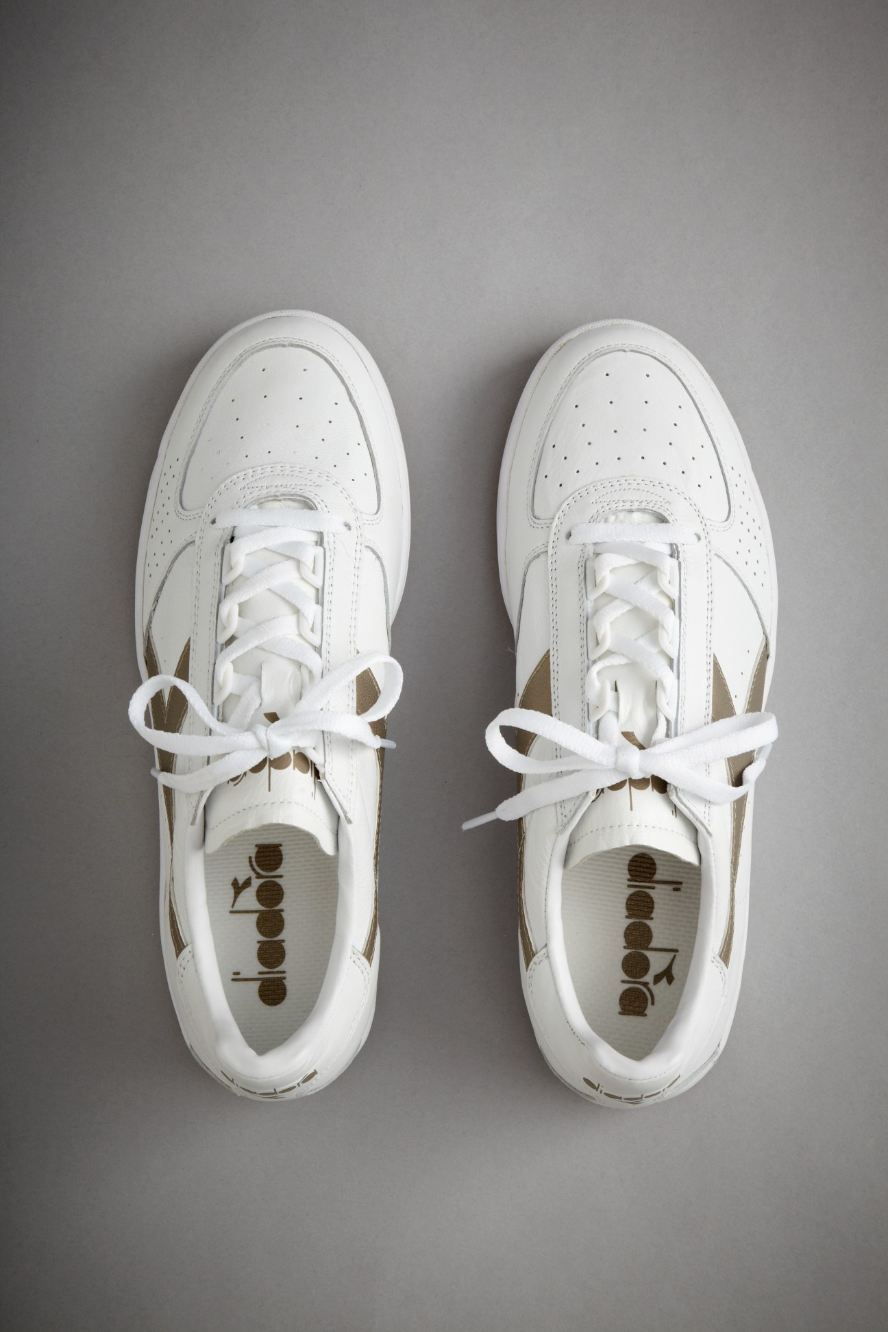Pair of white Diadora sneakers with perforated leather uppers and white laces on grey background.