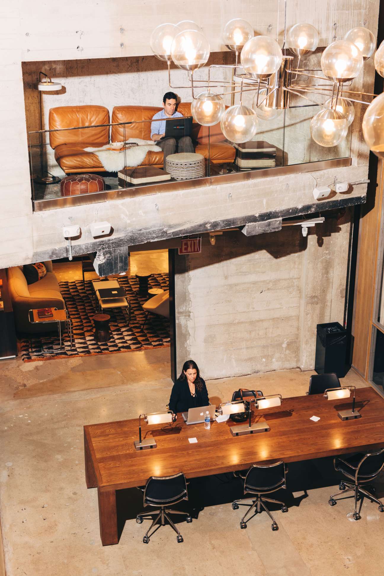 Multi-level co-working space at NeueHouse LA with woman at desk below and man on orange sofa above.