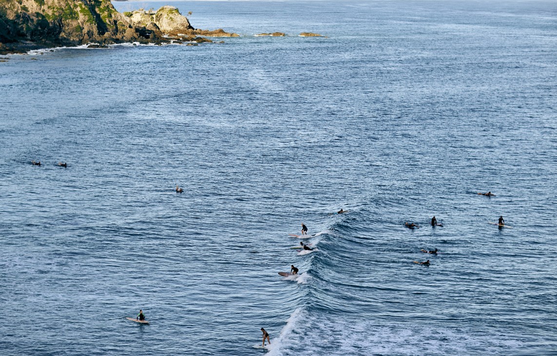 Surfers in wetsuits paddle out and ride waves at Wategos Beach with rocky headland in background.
