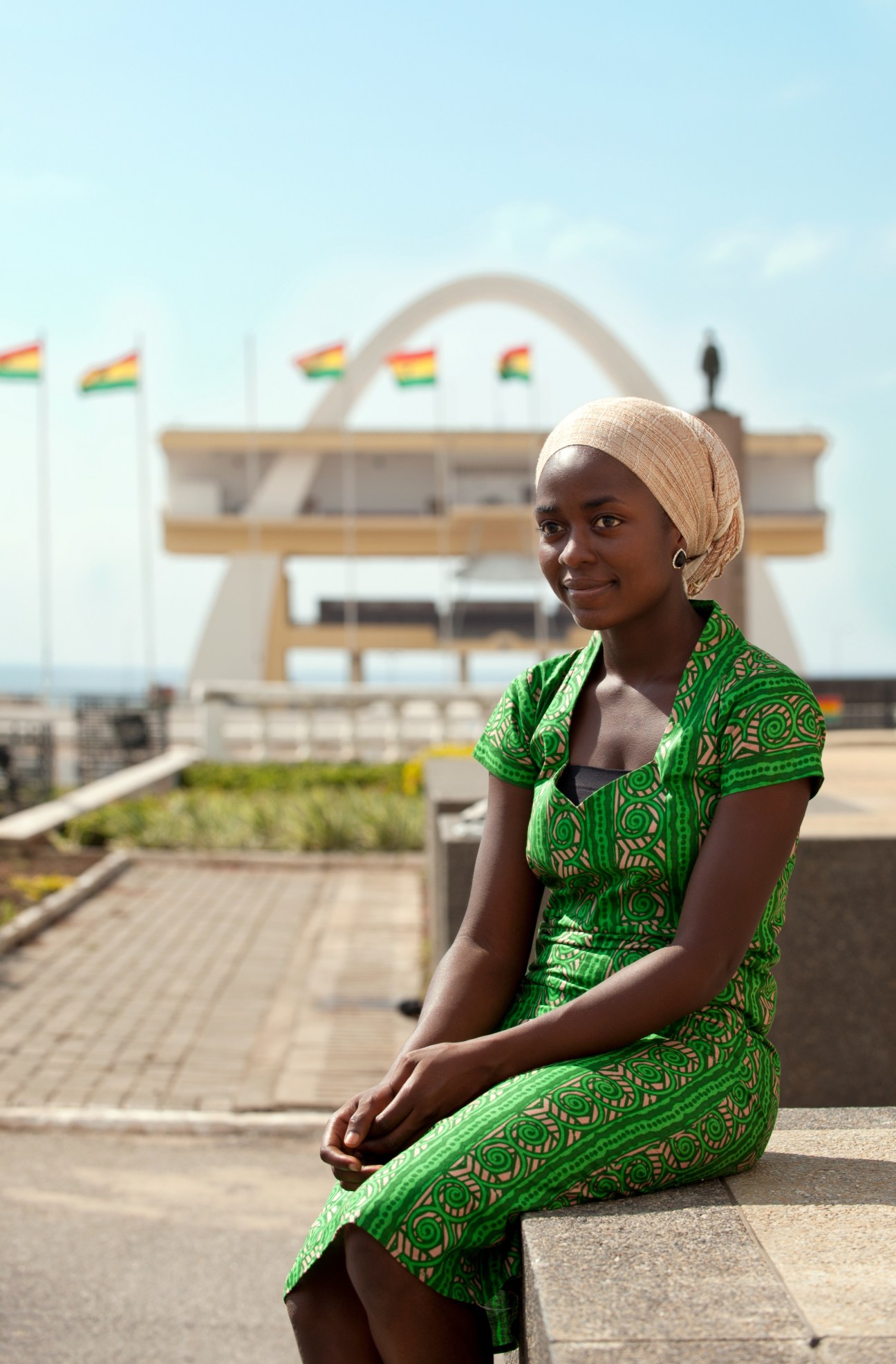Woman in green patterned dress smiling at camera with Ghana's Parliament House and flags in background.