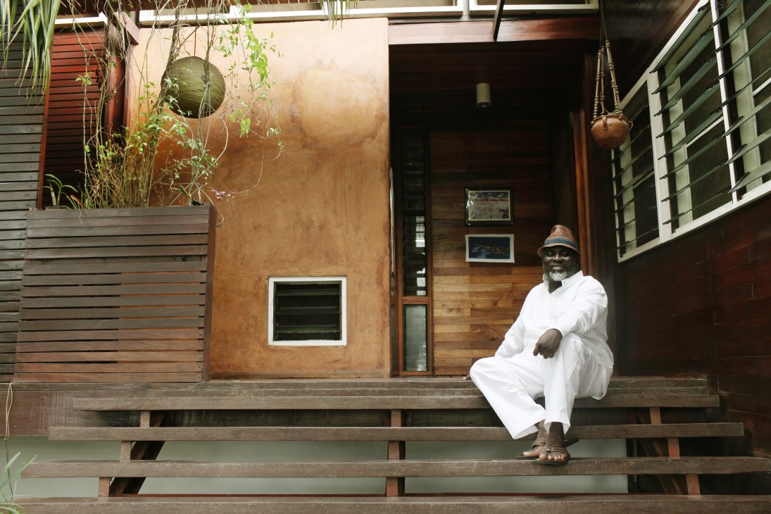 Architect Joe Addo sits on the wooden steps of his modernist house in Accra, wearing white clothing and a hat.