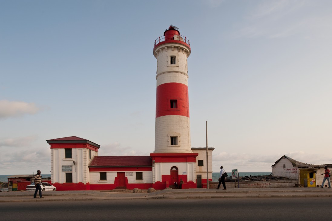 Red and white striped lighthouse with attached buildings on a beach in Jamestown, Ghana.