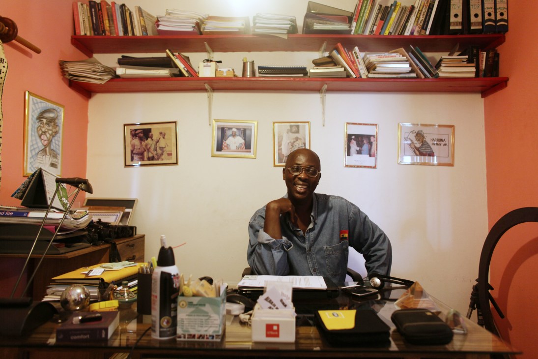 Harruna Attah, editor of Accra Mail, sits at his desk in his office with red shelves and framed photographs on the wall behind him.