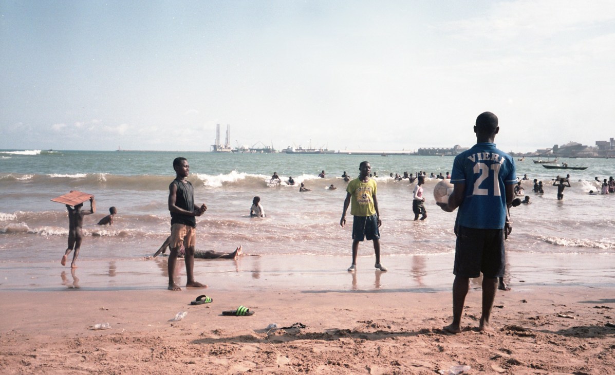 Children playing football on a beach in Takoradi with industrial port infrastructure visible on the horizon.